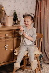 A little cute girl in gray pajamas with bows on her head sits on a chair near a wooden chest of...