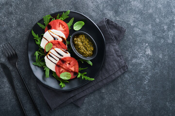 Caprese salad. Italian caprese salad with sliced tomatoes, mozzarella cheese, arugula, basil, olive oil in black plate over old brick tiles black background. Delicious Italian food. Top view.