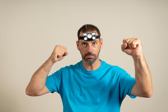 A Bearded Man With A Headlamp In A Hat Raising His Fists In Ostility, Isolated On Beige Background