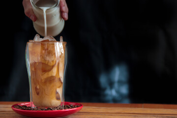 Ice coffee in a tall glass and pouring milk from above with coffee beans on the table in red plate.
