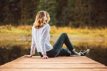 A beautiful blonde woman dressed in a white turtleneck and jeans is sitting in white headphones on a wooden pier near a forest lake on a summer evening. Relaxing with music in nature. Rest.