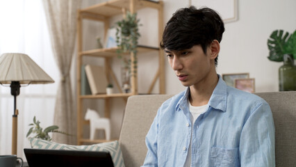 Fototapeta premium concentrated asian Japanese man frowning with a thoughtful face while working from home on the computer in the living room at daytime
