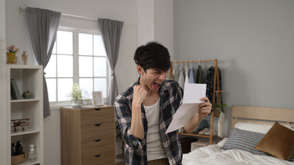 thrilled asian guy unfolding a paper letter is punching air with clenched fist and screaming with excitement for getting great test result in the bedroom at home - obrazy, fototapety, plakaty
