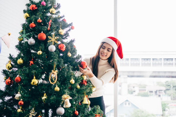 Happy young woman wearing a Santa Claus hat and sweater, smile and enjoy decorating various baubles on the Christmas tree in the living room at home. Enjoying and celebrating the Christmas holiday.