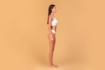 Half-turned slim woman in white underwear posing isolated on beige studio background, side view,...