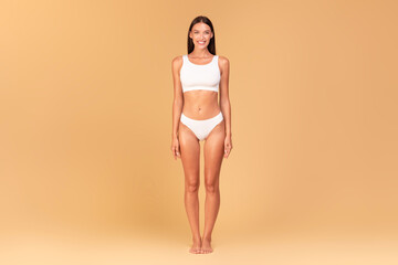 Full length frontal view of slim woman with perfect body shape and flat tummy posing on beige...