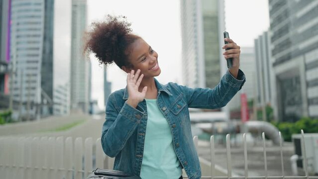Young tourist female talking to friends on mobile phone while standing outside on city street. Video call waving hand with modern skyscrapers background.