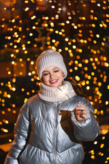 Girl with sparklers and Christmas garland. Smiling girl in warm clothes at night with blurry lights. Holidays theme