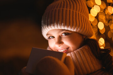 Girl and gift box with Christmas lights, close up. Smiling girl a light cap, scarf at night with blurry lights. Holidays theme