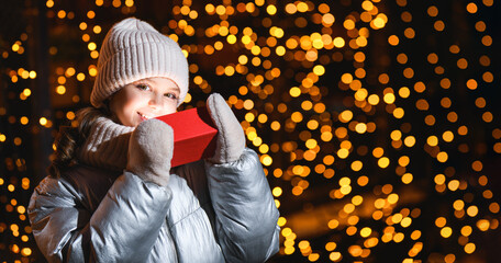 Girl and red gift box with Christmas lights. Smiling girl in a hat, scarf looking at the camera at night with blurry lights, copy space. Banner