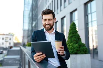 Smiling confident young caucasian guy with beard in suit has video call at tablet, hold cup of takeaway coffee