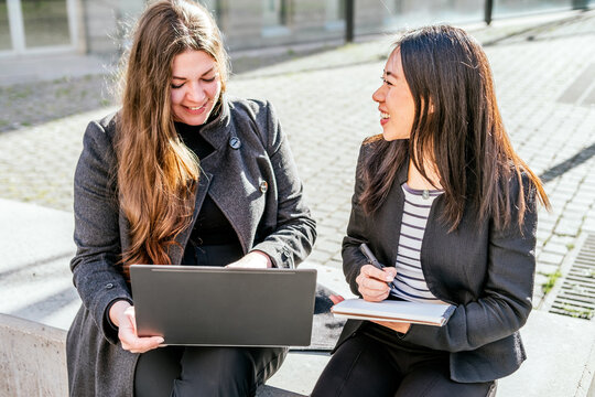 Positive woman typing on laptop while sitting near Asian female colleague writing in notepad while doing work together