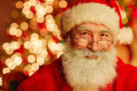 Close up portrait of traditional Santa Claus looking at camera and smiling with twinkling Christmas lights in background, copy space