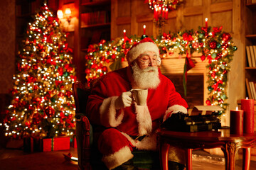 Portrait of traditional Santa Claus using typewriter and enjoying hot choco on Christmas eve