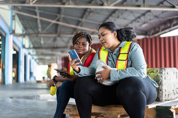 Two female African workers take a break, sitting and watching on mobile phone. chatting, sending...