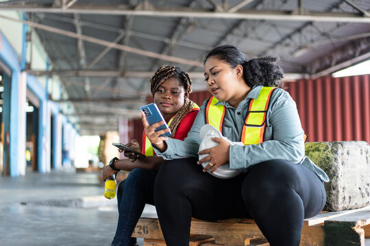 Two female African workers take a break, sitting and watching on mobile phone. chatting, sending message and browsing the internet at industrial factory