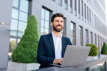 Cheerful confident handsome young caucasian guy with beard in suit typing on laptop