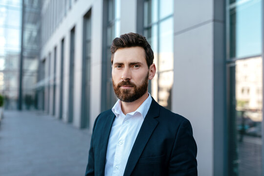 Headshot of serious attractive confident young caucasian guy with beard in suit looks at camera on street