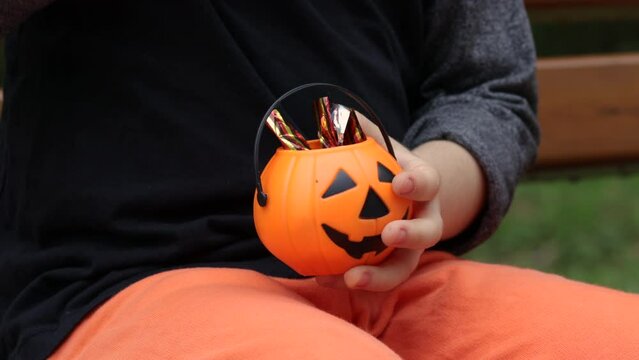 small cute halloween pumpkin bucket in kid little boy hand.child is smiling funny spooky.orange color bucket filled candies sweets wrapped in foil.kid hand take.park bench autumn fall trick or treat