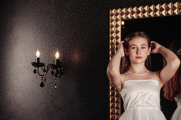 Portrait cute teen girl in an elegant white dress at mirror in dark stylish interior of living room. Teenager standing and posing, looking at camera. Concept of style, fashion and beauty. Copy space
