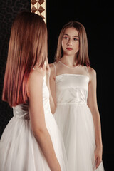 Fototapeta na wymiar Portrait cute teen girl in an elegant white dress at mirror in dark stylish interior of living room. Teenager standing and posing, looking at reflection. Concept of style, fashion and beauty
