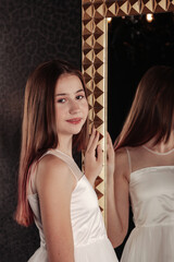 Teenager standing and posing, looking at camera. Portrait cute teen girl in an elegant white dress at mirror in dark stylish interior of living room. Concept of style, fashion and beauty. Copy space