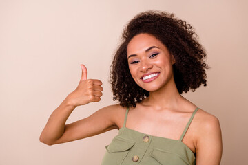 Photo of toothy beaming cheerful girl with curly hairstyle wear khaki overall showing thumb up like isolated on beige color background