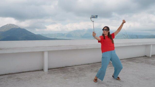a funny crazy blogger girl filming herself using stabilizer and cellphone camera as she is dancing on a terrace with Mountain View. Female traveler shooting funky dance movements using mobile phone.
