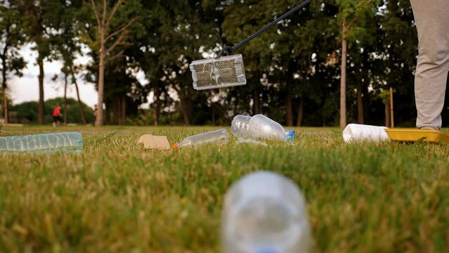 Close-up of a female activist collecting plastic garbage with tongs on the green grass. Volunteers help to save nature from plastic waste pollution.