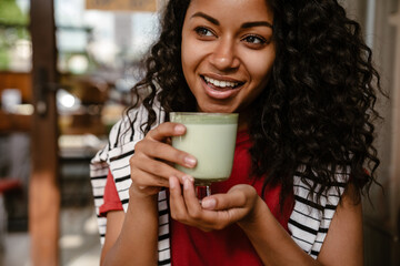 Young black woman smiling and drinking matcha latte at cafe