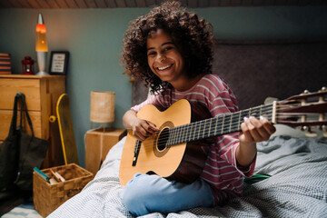 Black girl laughing and playing guitar while sitting on bed