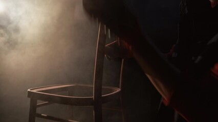 Furniture renovation. Man hobby. Woodwork renew. Carpenter sanding wood chair using sandpaper in dark home workshop with smoke cloud. - Powered by Adobe