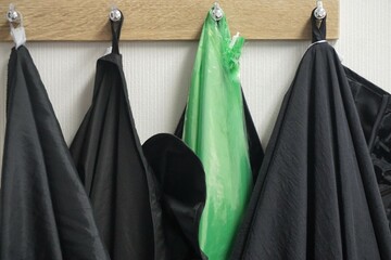 Schwarze Frisörkittel und grüne Plastikfolie hängen an Holzgarderobe vor weißer Wand in...