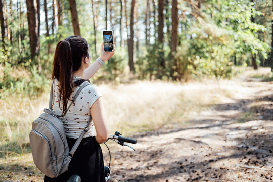 Young woman with backpack riding bike and taking photo on cell phone on pine forest background. Girl with bike shooting photo by phone camera in forest