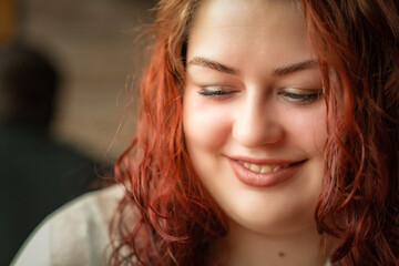 Naklejka na ściany i meble Close-up portrait of a young beautiful red-haired plump girl in a cafe.