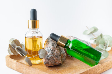 Naklejka premium glass dropper bottles with a pippette with gold rubber tip and green eucalyptus leaves near it on wooden tray on white background. Nature Skin concept. Organic Spa Cosmetics.