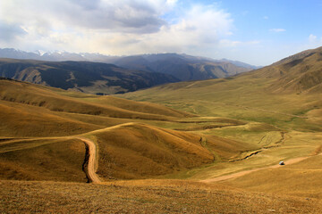 Top view of the Assy plateau, hills and slopes are covered with yellow grass and forest, long roads, a car is driving along the road, mountain ranges with snow-capped peaks in the distance, sky with c