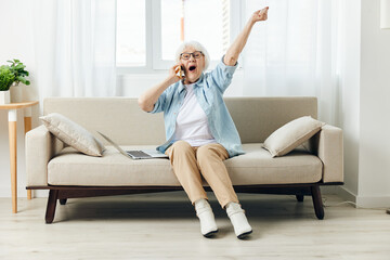 Fototapeta na wymiar an enthusiastic, emotional elderly woman is sitting on the sofa in an apartment and shouts widely with happiness, raising her hand up during a telephone conversation