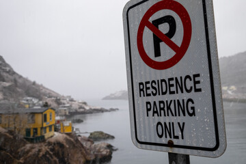 A white metal sign with residence parking only on a metal post. The sign overlooks a foggy harbour with colorful houses situated on a hillside. There's a narrow entrance to St. John's Harbour. 