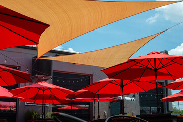 Multiple triangle shaped yellow nylon sunshades and awnings hanging over a patio deck. There are red colored canvas umbrellas hung with strings of clear patio light against a bright blue sunny sky. - obrazy, fototapety, plakaty