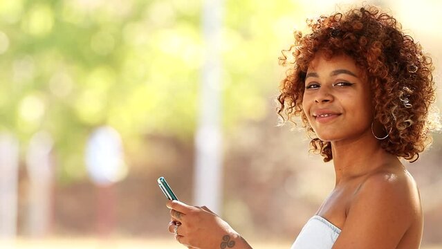 latin afro american young woman smiling looking at camera with mobile phone outdoors
