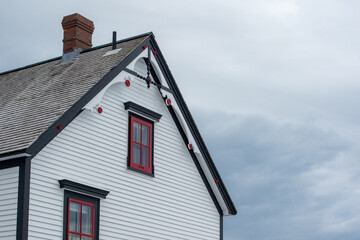 Exterior white wooden wall of horizontal clapboard siding on a vintage house with black trim. There are small four pane glass windows with red trim and a decorative gable cedar shake roof.