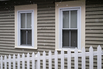 White picket fence on a diagonal in the foreground with a tan coloured house in the background. There are two white double hung windows with beige trim on the exterior wall of the country style home. 