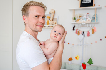 Obraz na płótnie Canvas A young caucasian father gently holds a baby in a bright room at home. Happy fatherhood and the joy of being a father