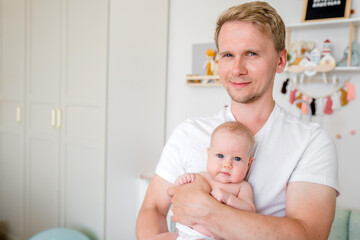 A young caucasian father gently holds a baby in a bright room at home. Happy fatherhood and the joy...