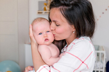A young Caucasian mother gently hugs a baby in a bright room for a baby at home. Motherhood and happiness, emotions.
