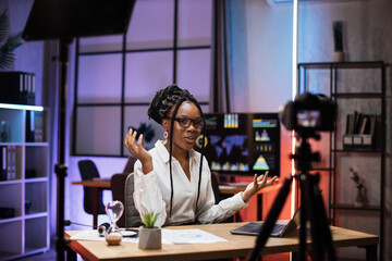 Confident african american businesswoman, broker in white shirt sitting in front of camera in evening office during recording video for business vlog.