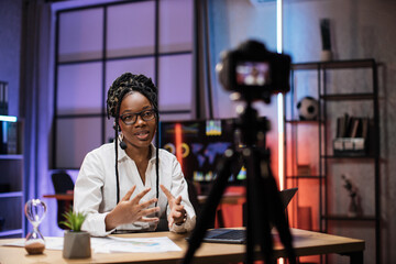 Portrait of attractive confident experienced smart female african american office manager in headset recording video vlog for internet audience in evening office.