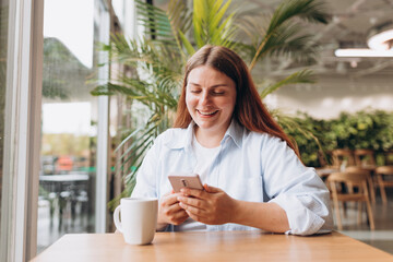 White brunete woman using smartphone while sitting in cafe indoors. Young woman at cafe drinking coffee or tea indoors on a rainy day. Concept of easy breakfast.