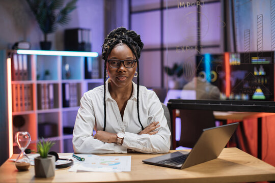 Front view portrait of young smiling experienced smart stylish african american female office manager in white shirt sitting at table using laptop in evening office.